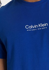 Niebieska bawełniana koszulka z okrągłym dekoltem, krótkimi rękawami oraz białym nadrukiem z napisem "Calvin Klein" i współrzędnymi geograficznymi.