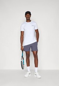 Muž v bílém tričku, námořnických šortkách, bílých ponožkách a botách, držící tenisovou raketu, stojící na jednoduchém světlém pozadí.