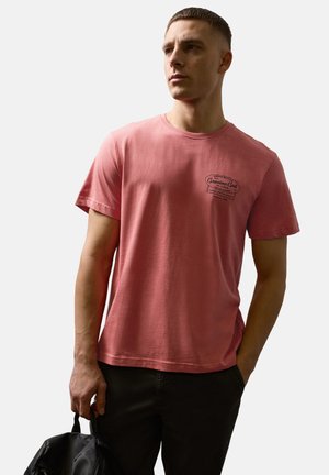 Junger Mann, der ein ausgewaschenes rotes T-Shirt mit Grafik und schwarze Hose trägt, eine schwarze Tasche in einer Hand hält und seitlich gegen einen einfarbigen Hintergrund schaut.