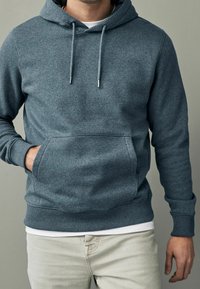 Sudadera con capucha gris, fabricada en un tejido suave, con cordones, bolsillo frontal, puños y dobladillo de punto. Combinándola con jeans de color claro.