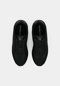 Pereche de adidași negri Calvin Klein Jeans cu șireturi, logo-ul vizibil pe limbă și pe lateral, văzuți dintr-o parte de sus pe un fundal alb.