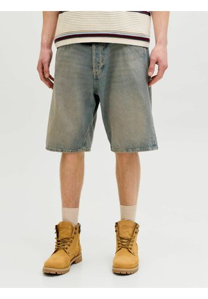 Muž v vybledlých modrých džínových šortkách, béžových ponožkách a hnědých pracovních botách na šněrování, zobrazený od poloviny stehna dolů na bílém pozadí.