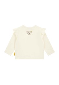 Long-sleeve cream top with ruffle accents on one shoulder, featuring an embroidered bear face patch on the back. Soft texture, casual design.