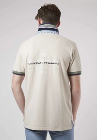 Polo beige avec un col à motifs, logo brodé au dos et accents rayés aux manches. Texture douce et coupe régulière.