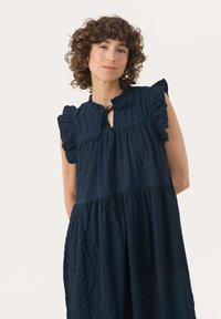 Navy sininen mekko, joka on valmistettu tekstuurikankaasta, jossa on röyhelöhihoja, avainaukko-olkaimet ja kerroksellinen siluetti.