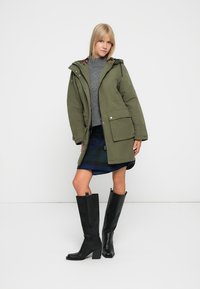 Oliven grøn parka med aftagelig hætte, frontlommer, lagdelt over en grå sweater og rutet nederdel, parret med høje sorte hælebluser.