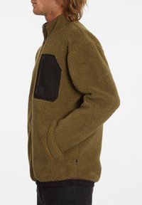 Fleecejacka i olivgrönt med en svart bröstficka med dragkedja. Har en ståkrage, mjuk textur och sidofickor.