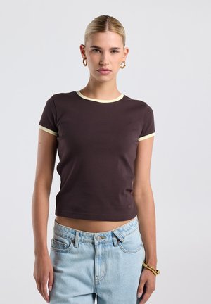 Jonge vrouw met blond haar die een donkerbruin cropped T-shirt met lichte biezen en lichtblauwe high-waisted jeans draagt, staand tegen een witte achtergrond.