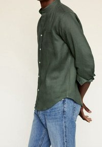 Camisa de lino verde oscuro con cuello mandarín, bolsillo delantero y cierre de botones, combinada con vaqueros de mezclilla azul claro.