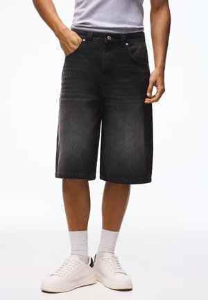 Mann trägt schwarze kniehohe Jeansshorts, weiße Socken und weiße Turnschuhe, steht mit einer Hand in der Tasche.