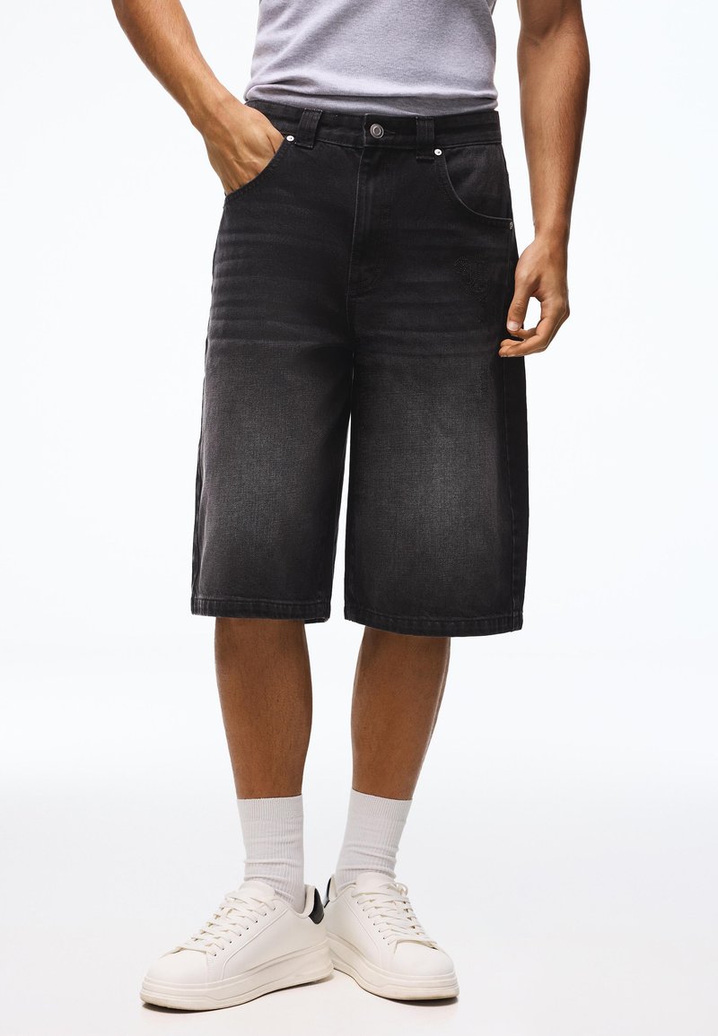 Homme portant un short en denim noir longueur genou, chaussettes blanches et baskets blanches, debout avec une main dans la poche.