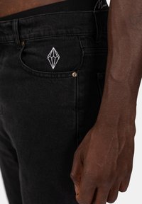 Svarta denimjeans har vit rombformig broderi på främre fickan och en borstad textur. Midjebandet är synligt ovanför byxorna.