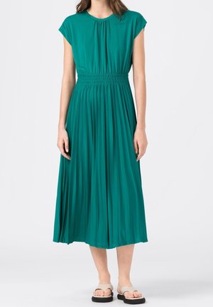 Woman wearing a short-sleeve teal midi dress with a gathered waist and pleated skirt, paired with beige platform sandals.