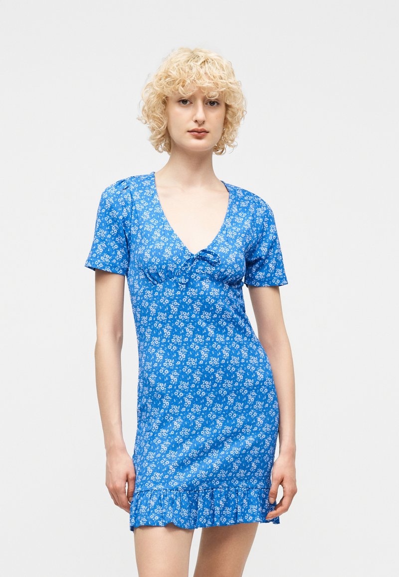 Νέος με σγουρά ξανθά μαλλιά, ντυμένος με κοντό μπλε φόρεμα με λουλούδια, κοντά μανίκια και βολάν στο τελείωμα, στέκεται μπροστά σε ένα απλό φόντο.