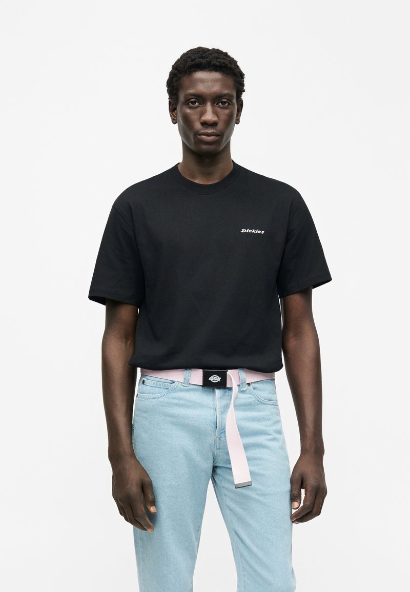 Camiseta negra de algodón con mangas cortas, con un logo blanco. Combinada con vaqueros de mezclilla azul claro y un cinturón rosa, creando un look casual.