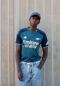 Maglia da calcio teal con accenti bianchi e blu, con la scritta "Emirates Fly Better", abbinata a jeans azzurri e un cappellino blu.