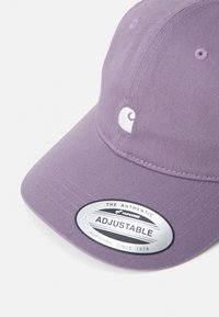 Lilla cap laget av bomull, med justerbar stropp, avrundet krone, og en hvit brodert logo på fronten. Sølv klistremerke på skjermen.