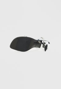 Ασημένια σανδάλια με τακούνι που διαθέτουν ένα κομψό, διαφανές λουράκι και έναν απαλό μαύρο πάτο με καμπύλο τακούνι και στρογγυλή μύτη.