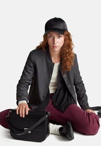 Černá bomber bunda, bílý žebrovaný top, burgundské kalhoty, černá kožená čepice s logem, texturovaná černá kabelka, černé boty.