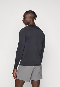 Μακρυμάνικη αθλητική μπλούζα σε μαύρο χρώμα με λεία υφή, στρογγυλή λαιμόκοψη και διακριτικές λεπτομέρειες ραφής. Φοριέται με γκρι σορτς.