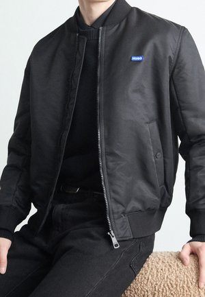 Homme portant une veste bomber noire zippée sur un pull noir, assis sur une surface beige texturée, cadré au niveau du cou et des cuisses.