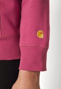 Roze katoenen sweatshirt met ribgebreide manchetten, afgewerkt met een gele geborduurde logo op de elleboog. Zachte textuur en een casual ontwerp.