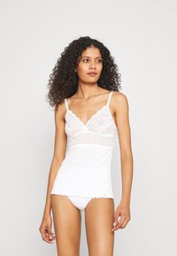 White camisole with lace trim and cut-out detailing, adjustable straps, and a fitted silhouette. Soft, smooth fabric with a feminine design.