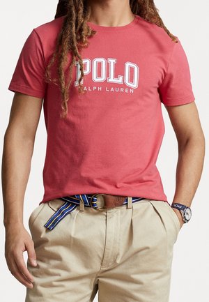 Osoba nosząca czerwoną koszulkę Polo Ralph Lauren, beżowe spodnie z niebieskim paskiem w paski i zegarek na lewej ręce.
