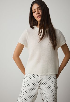 Donna con capelli lunghi e scuri che indossa un top bianco a maniche corte in maglia e pantaloni bianchi con pois neri, in piedi con le mani dietro la schiena.