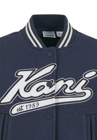 Jūras zilās universitātes jakas ar baltu strīpētu apkakli, pogām ar snap ievilkumu un drosmīgu "Kari" logo ar "est. 1989" detaļu. Gluda auduma virsma.
