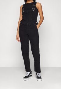 Zwarte overalls van denim met verstelbare bandjes, rechte pijpen, een voorzak en zijzakken, gecombineerd met zwarte hoge sneakers.