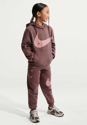 Joven chica sonriendo, vistiendo una sudadera con capucha Nike color malva y pantalones deportivos con logos swoosh rosas, combinados con zapatillas blancas y malva, de pie con las manos en los bolsillos.