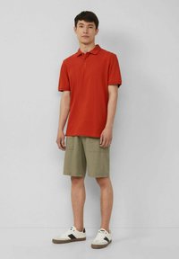 Rotes Polohemd mit Kragen, kombiniert mit olivgrünen Cargo-Shorts und weißen Sneakers mit braunen Akzenten.