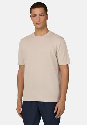 Jeune homme aux cheveux courts et foncés, portant une chemise beige à manches courtes et un pantalon bleu foncé, debout devant un fond clair uni.