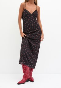 Robe nuisette noire à imprimé floral, fines bretelles et bordure en dentelle. Le mannequin porte des bottes de cowboy rouges à bouts pointus avec des détails de couture blanche élaborée.