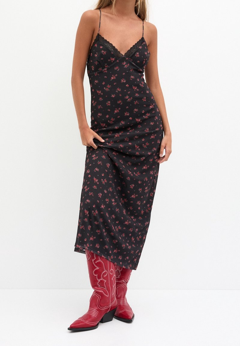 Robe nuisette noire à imprimé floral, fines bretelles et bordure en dentelle. Le mannequin porte des bottes de cowboy rouges à bouts pointus avec des détails de couture blanche élaborée.