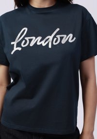 Camiseta de algodón de color verde oscuro con un grande texto en glitter plateado que dice "London" en un estilo de escritura cursiva en la parte frontal. Mangas cortas y cuello redondo.