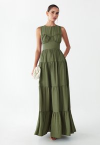 Vrouw draagt mouwloze olijfgroene maxijurk met lagen, houdt witte gevlochten clutch vast, staat voor een witte achtergrond.