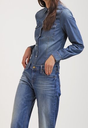 Vrouw die een nauwsluitend blauw denimhemd draagt met lange mouwen en hoge blauwe spijkerbroek met lichte vervaging, staande met handen dicht bij de zakken.