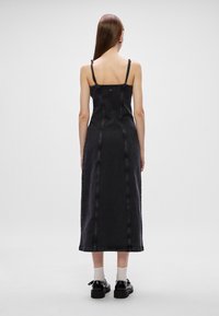 Vestido maxi de denim preto com alças finas, apresentando um corpete ajustado e costuras visíveis. Usado com sapatos pretos e meias brancas.