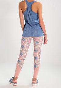 Débardeur de sport bleu avec un design dos nageur, associé à un legging à motifs présentant des tons corail, rose et bleu avec des designs abstraits. Baskets grises.