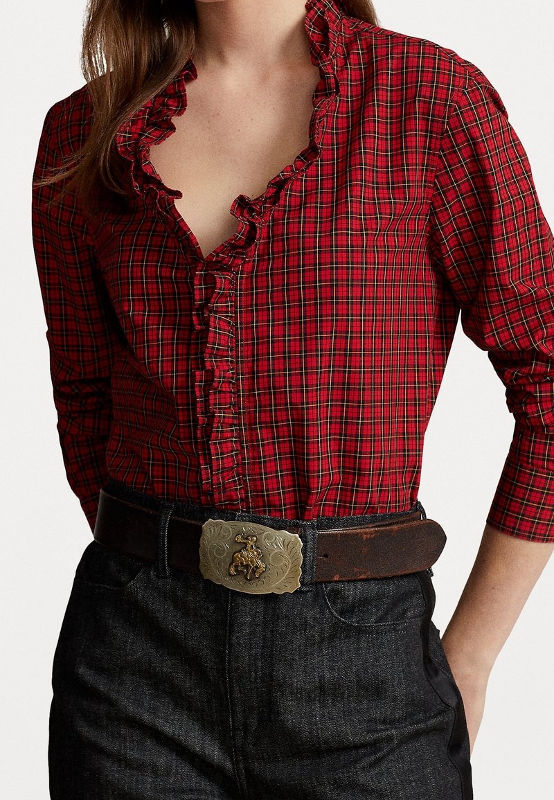 Femme portant un chemisier à carreaux rouges avec un col froufroutant rentré dans un jean noir et une ceinture en cuir marron avec une boucle métallique ornée.