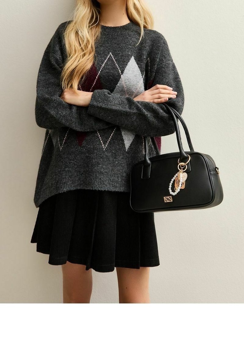 Femme en pull gris à losanges et jupe noire plissée tenant un sac à main noir avec un pendentif en or et perles devant un mur uni.