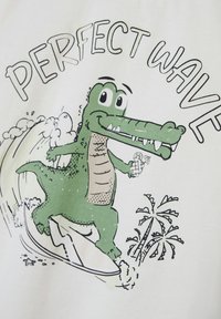 White T-shirt with a cartoon crocodile surfing, holding a drink. Features green and black colors, with text "PERFECT WAVE" above the image.
