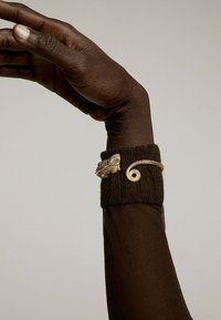Brazalete con acentos dorados sobre un puño de punto marrón, que presenta un diseño de hoja y un patrón en espiral, llevado en una mano de piel oscura y levantada.