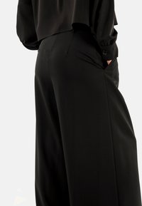 Pantaloni neri a gamba larga realizzati in tessuto morbido, dotati di tasche laterali e vestibilità su misura, con un accento a bottone sui polsini di un top stratificato.
