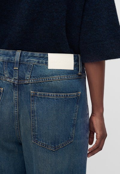 Gros plan d'une personne portant un jean en denim bleu avec une étiquette en cuir blanche et un pull bleu marine foncé, montrant la poche arrière et la main droite.