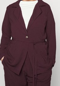 Blazer texturizado burdeos con un solo botón, cuello con muesca y cinta en la cintura, combinado con una blusa blanca que cuenta con dos bolsillos frontales.