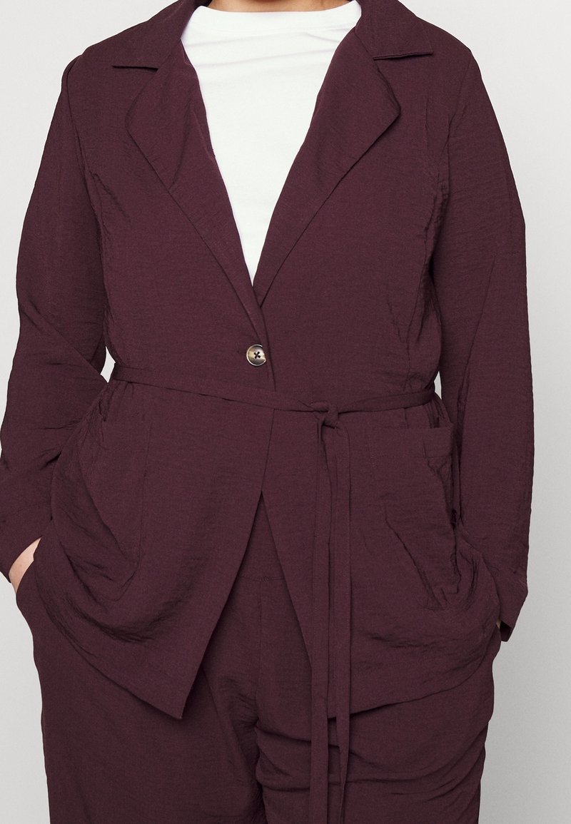 Blazer texturizado burdeos con un solo botón, cuello con muesca y cinta en la cintura, combinado con una blusa blanca que cuenta con dos bolsillos frontales.