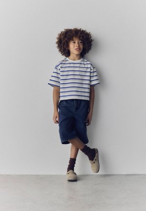 Child with curly hair wearing a striped t-shirt, denim shorts, brown socks, and beige shoes, standing against a plain light gray wall.